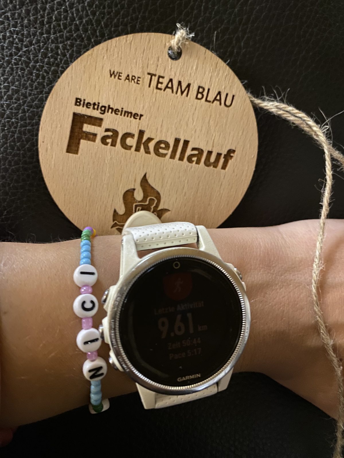 Eine Holzmedaille neben einer Laufuhr, am selben Handgelenk ein Perlenarmband mit den Buchstaben N I C I